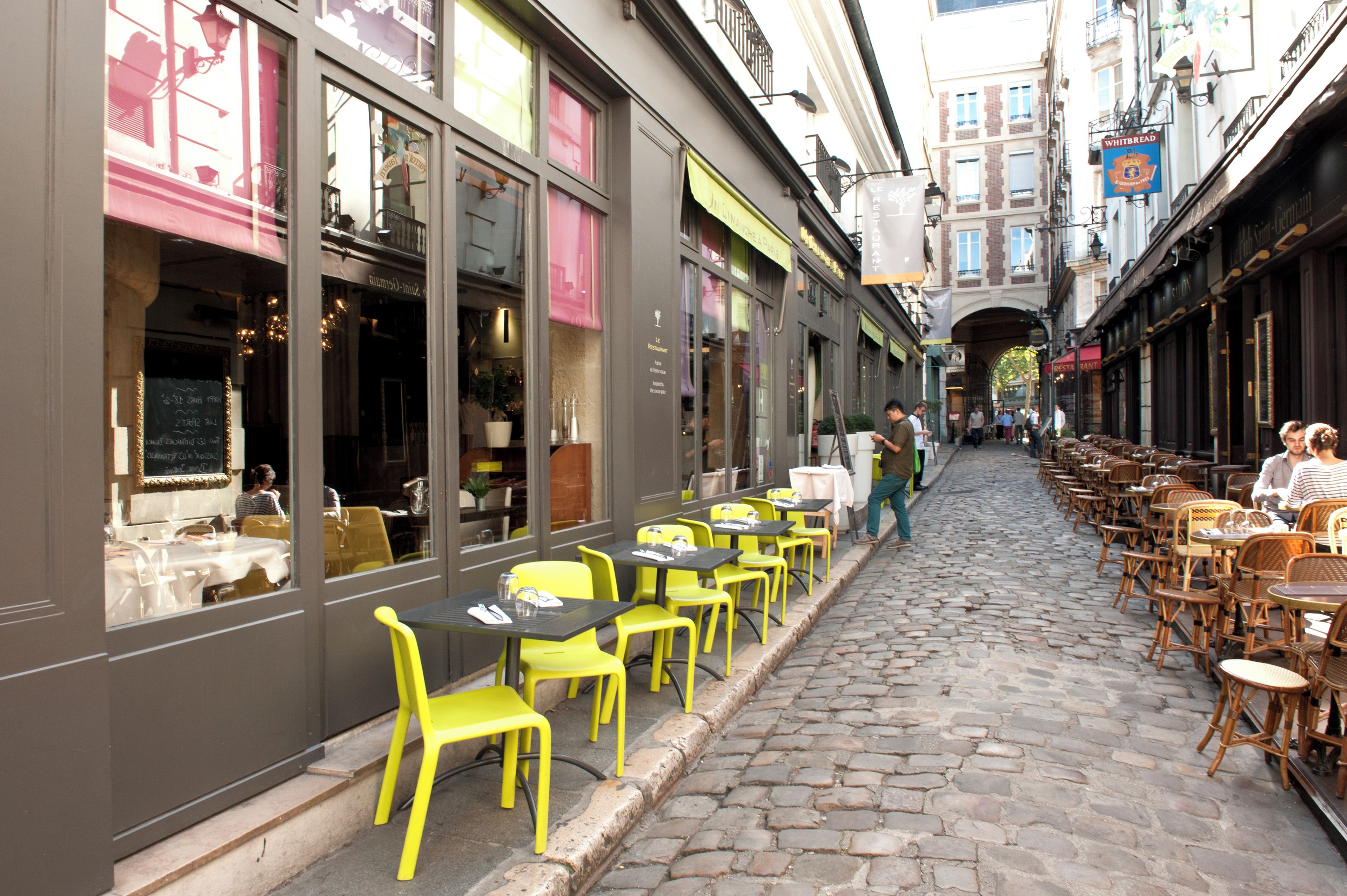 A cobblestone street with outdoor seating and tables, surrounded by buildings with large windows and signage.