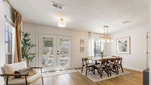 A spacious dining area with a wooden table and chairs, a sofa with a throw blanket, and a large window with curtains. - The Cousins House, private pool, prime location (Rogers)
