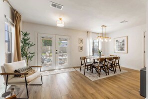 A spacious dining area with a wooden table and chairs, a sofa with a throw blanket, and a large window with curtains. - The Cousins House, private pool, prime location (Rogers)