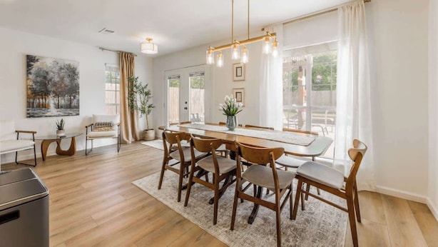 A modern dining area with a wooden table and chairs, a hanging light fixture, and a large window with curtains. - The Cousins House, private pool, prime location (Rogers)
