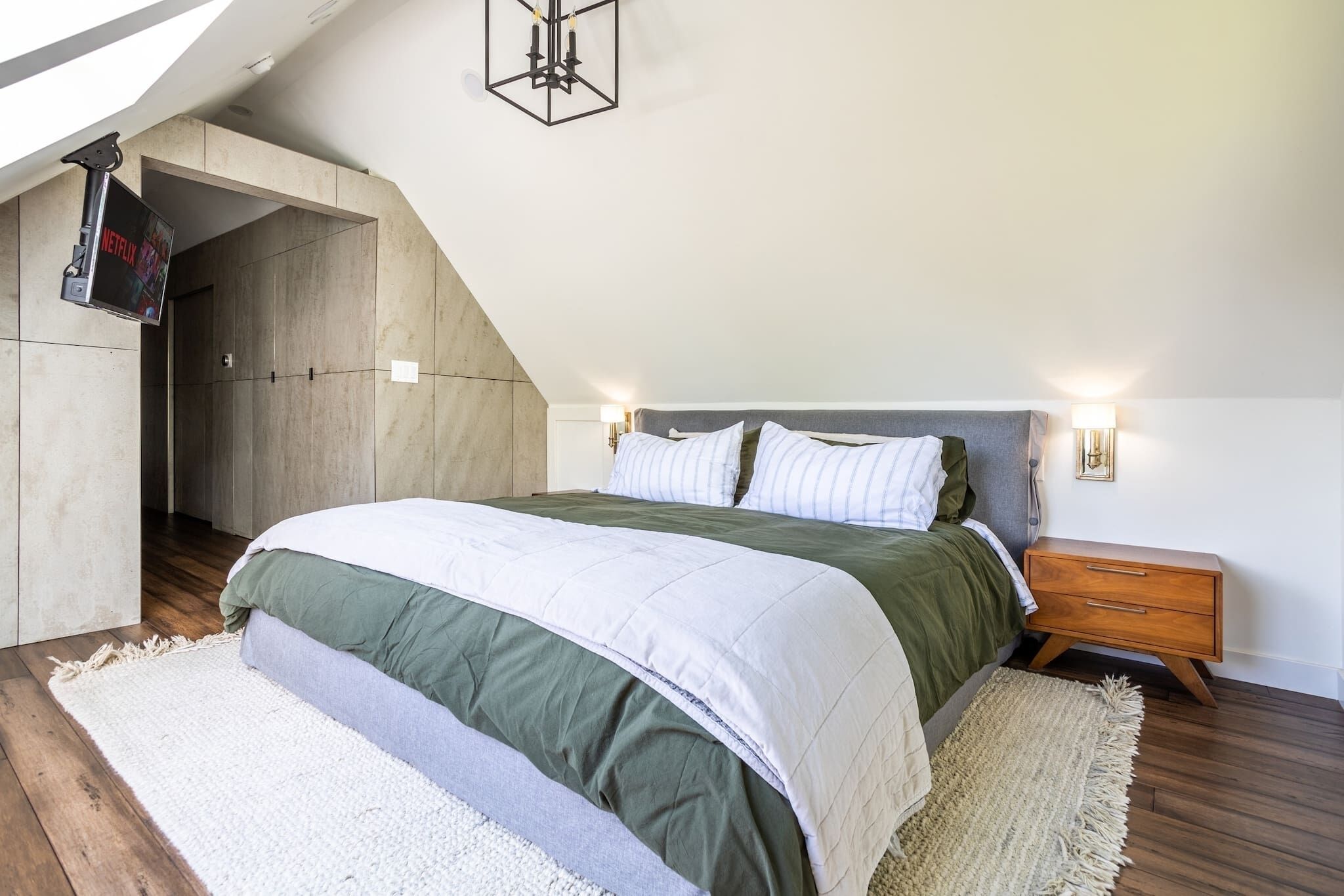 A bedroom with a sloped ceiling, a bed with green and white bedding, a wooden nightstand, and a wall-mounted TV.