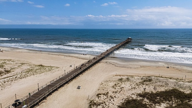 Una playa con un muelle largo que se adentra en el océano, dunas arenosas y un cielo despejado.