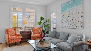 Un salón con un sofá gris, dos sillones estampados color naranja, una mesa de centro de vidrio y un cuadro abstracto grande.