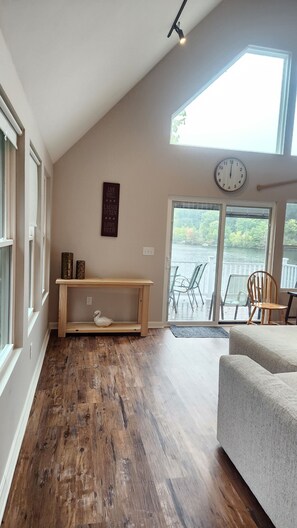 A room with a sloped ceiling, a wooden bench, a sofa, a clock, and a view of a lake through large windows. - Lakeside Paradise with Private Dock. (Weare)