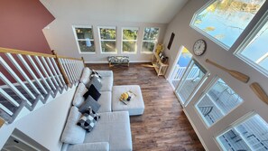 A spacious living area with a gray sectional sofa, wooden flooring, and multiple windows offering views of the outdoors. - Lakeside Paradise with Private Dock. (Weare)