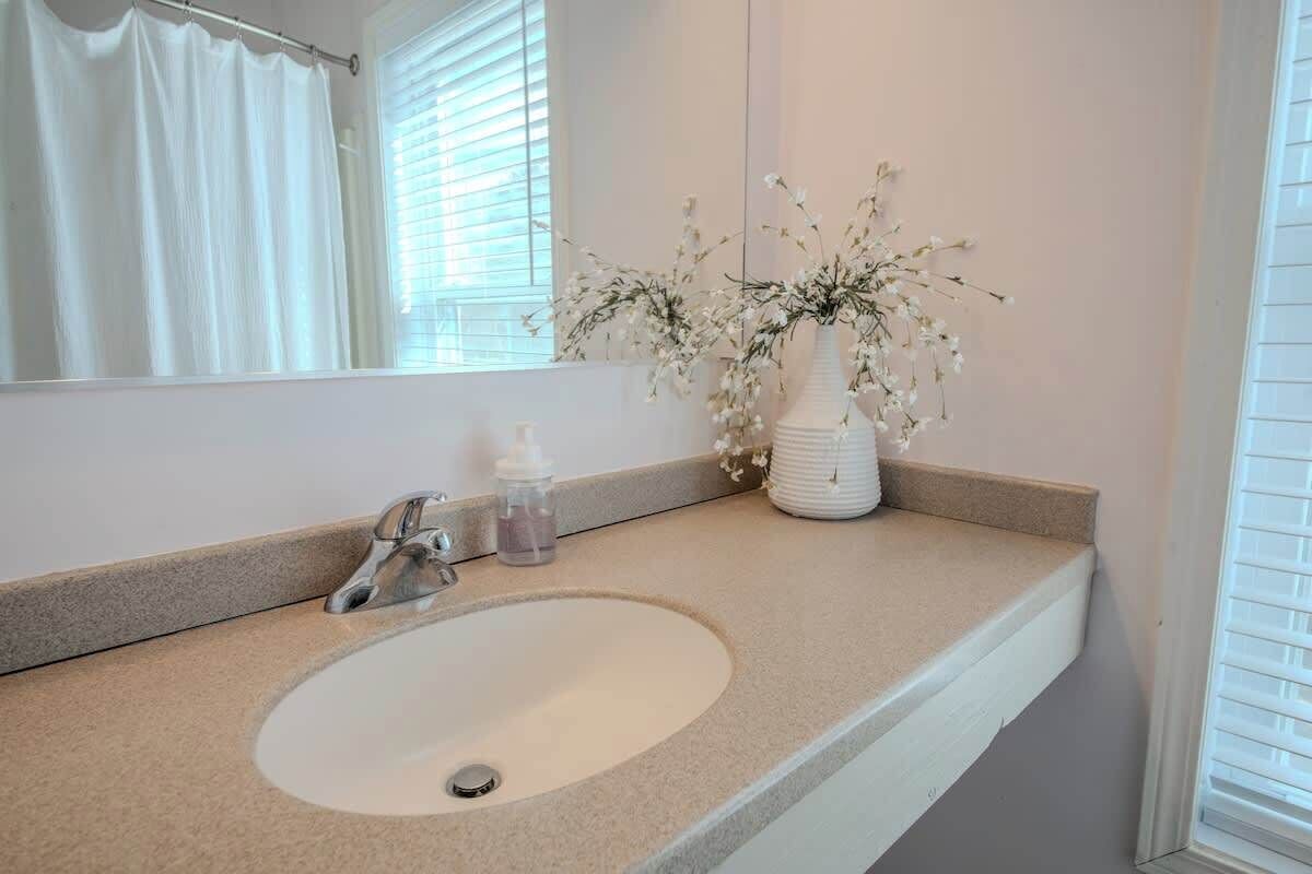 Une salle de bain avec un lavabo blanc, un miroir, un vase de fleurs et un rideau de douche.