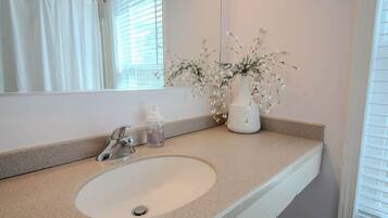Une salle de bain avec un lavabo blanc, un miroir, un vase de fleurs et un rideau de douche.