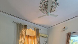 A bedroom with a bed, a desk, a chair, an air conditioning unit, and a window with curtains. - Hotel Pushkar Heritage (Pushkar)