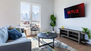 A modern living room with a grey sofa, a round coffee table, a black chair, a TV mounted on the wall, and a city view through a large window.