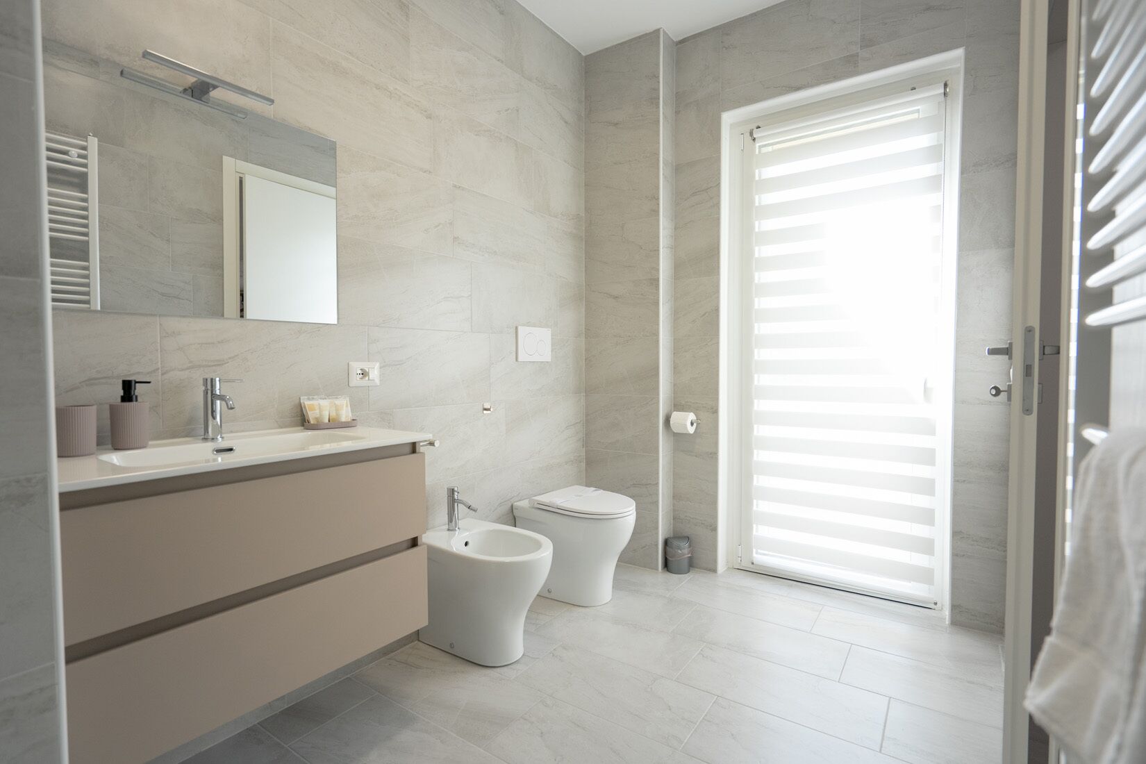 Un baño moderno con tocador, bidé, inodoro y un ventanal grande con persianas.