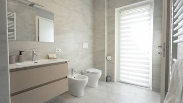 Un baño moderno con tocador, bidé, inodoro y un ventanal grande con persianas.