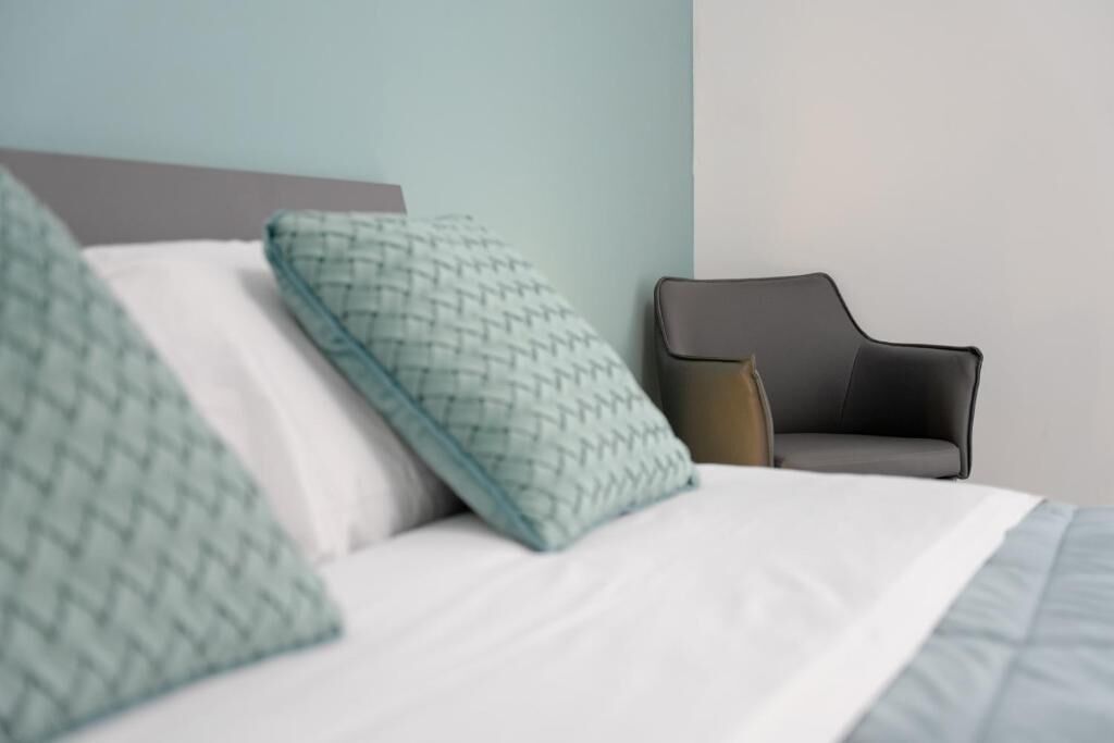 A bed with patterned pillows, a grey chair, and a plain wall.