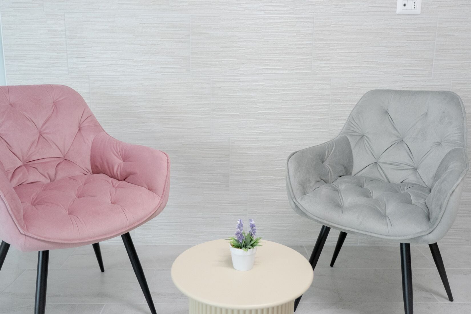 Dos sillones tapizados con respaldo capitonado y patas negras, una mesita redonda con superficie blanca y una planta en maceta con flores moradas.