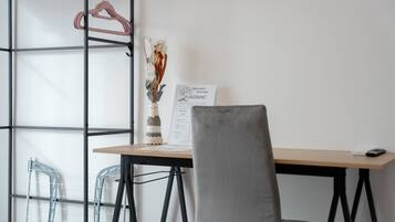 Una habitación con escritorio, una silla, un perchero con ropa y un jarrón con flores secas.