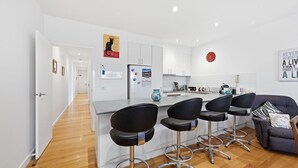 A modern kitchen with white cabinets, a refrigerator, and a dining area with bar stools. - Tide & Town - Perfectly Located! (Inverloch)