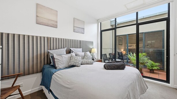 A modern bedroom with a large bed, striped headboard, and a view of the outdoors through large windows. - Tide & Town - Perfectly Located! (Inverloch)
