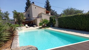 Una piscina rectangular con un muelle de madera al frente, una casa con techo inclinado al fondo y un seto a la derecha.
