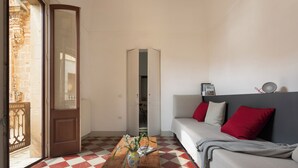 Ein Zimmer mit Schachbrettmuster-Boden, einem Holztisch mit einer Vase, einem Sofa mit roten Kissen und einer Tür, die zu einem Außenbereich führt.