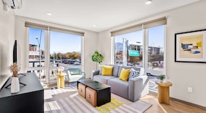 Un salon moderne avec un canapé, une table basse et des tables d’appoint, de grandes fenêtres offrant une vue sur la ville et un tableau encadré accroché au mur.