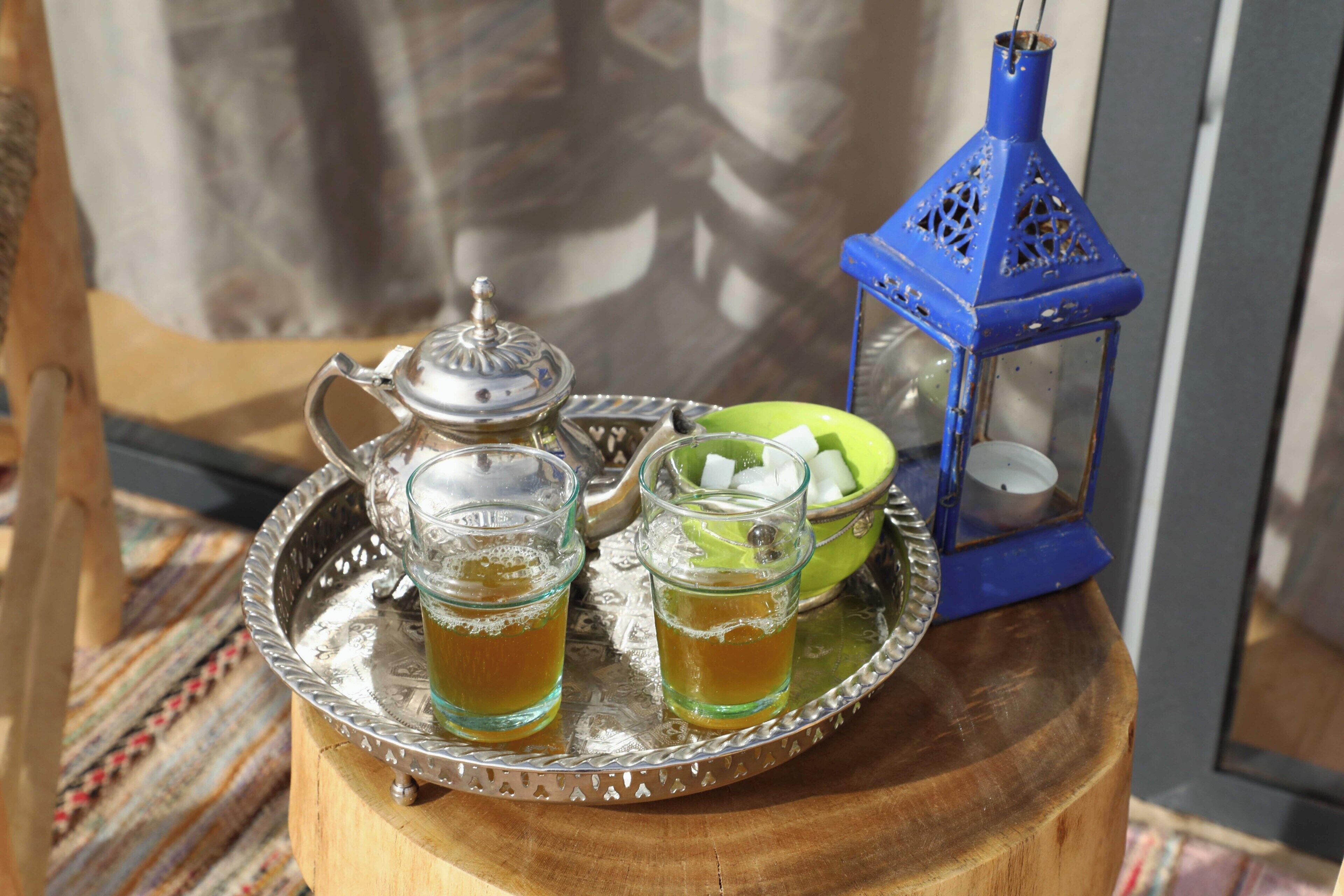 A tray with a teapot, two glasses of tea, a bowl of fruit, and a blue lantern on a wooden stand.