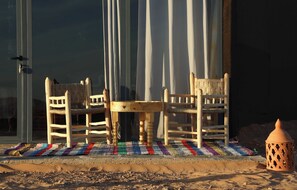 Une table et des chaises en bois, accompagnées d’un tapis tissé, d’un panier en osier et d’une lanterne, le tout posé sur une surface sablonneuse.