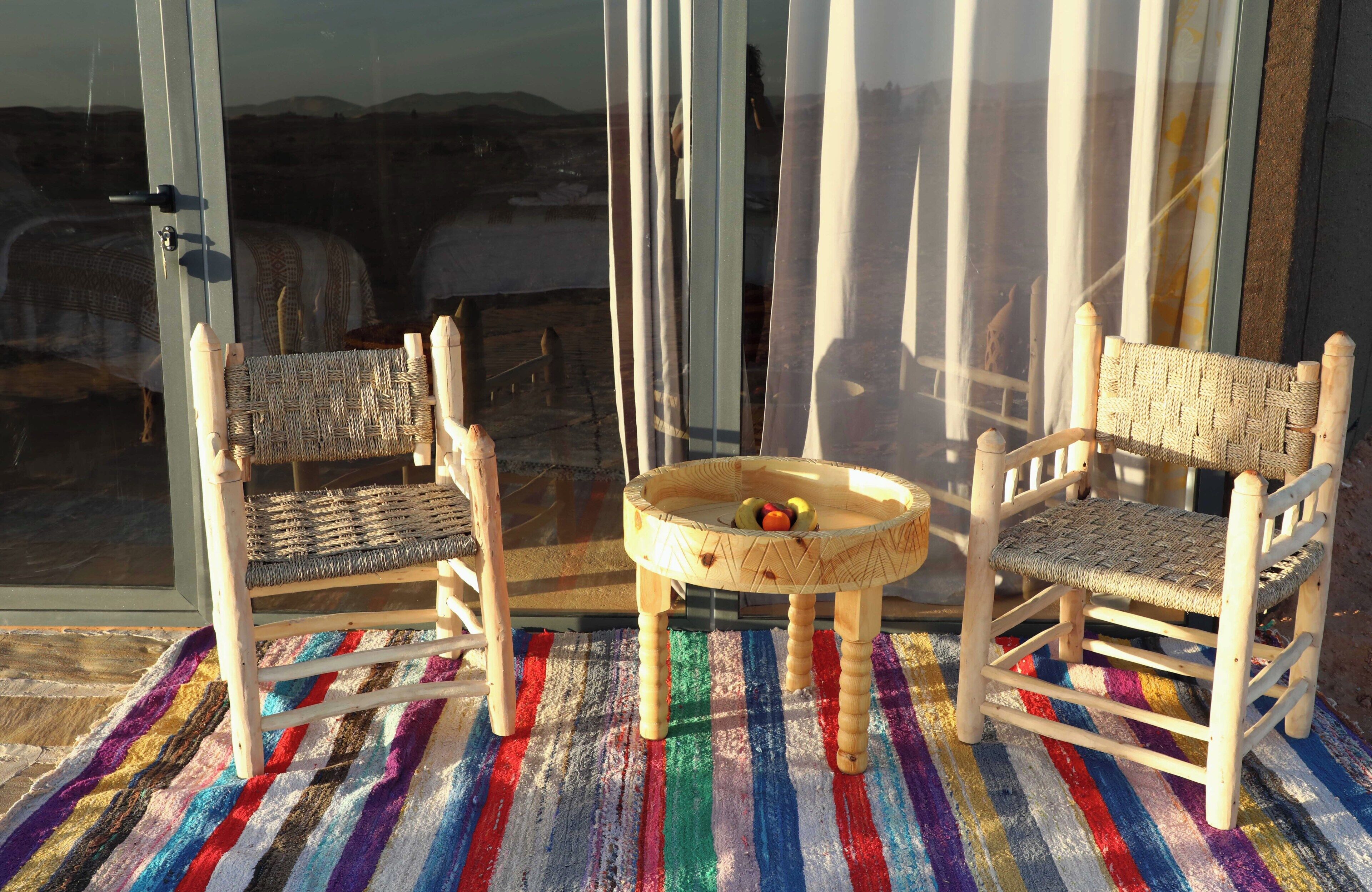 Two wooden chairs with woven seats and backs, a small round table with fruits, and a colorful striped rug on a balcony with a view of the landscape.