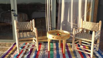 Two wooden chairs with woven seats and backs, a small round table with fruits, and a colorful striped rug on a balcony with a view of the landscape.