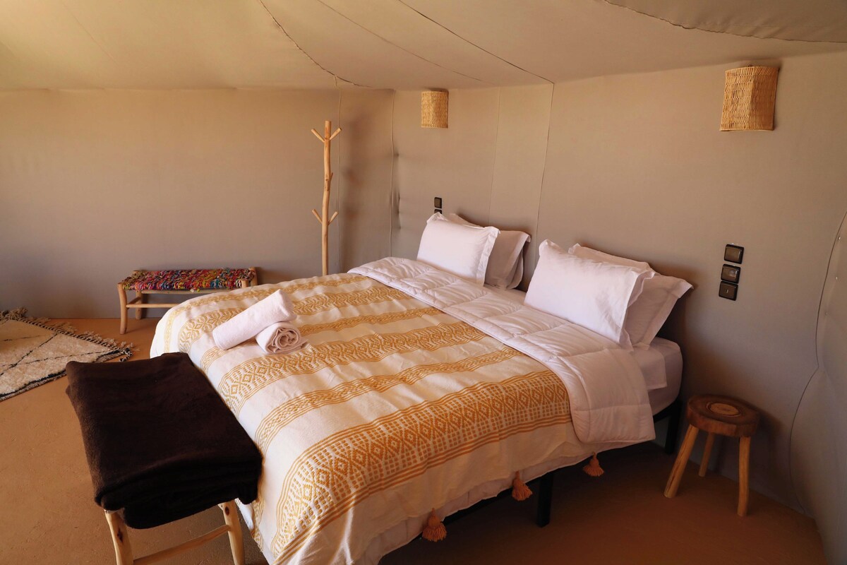 Ein Bett mit einem gemusterten Überwurf, eine Holzbank, ein kleiner Hocker und ein gewebter Wandbehang.