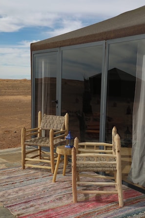 A wooden chair and table set outside a building with large glass doors, a woven rug on the ground, and a desert landscape in the background. - Hamada Retreat (Tisserdmine)