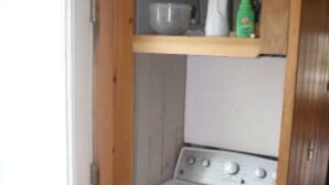 Pequeña lavandería con lavadora, estante superior y puerta a la izquierda.