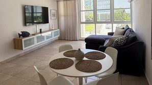 Een moderne woonkamer met een eethoek, grote ramen en een flatscreen televisie aan de muur.