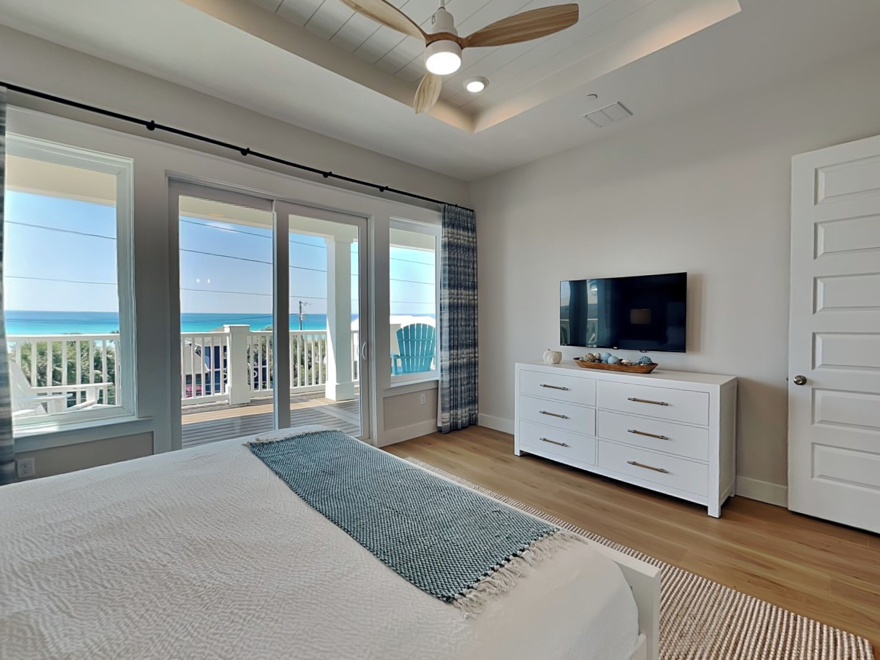 A bedroom with a large bed, a TV on a white dresser, and a balcony with a view of the ocean.