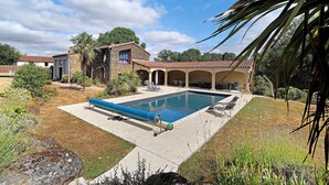 A swimming pool with a blue cover, a stone house with a red door, and a covered patio area. - Petite house “La Grande Rivière” with pool, Wi-Fi, and air conditioning (Sèvremoine)