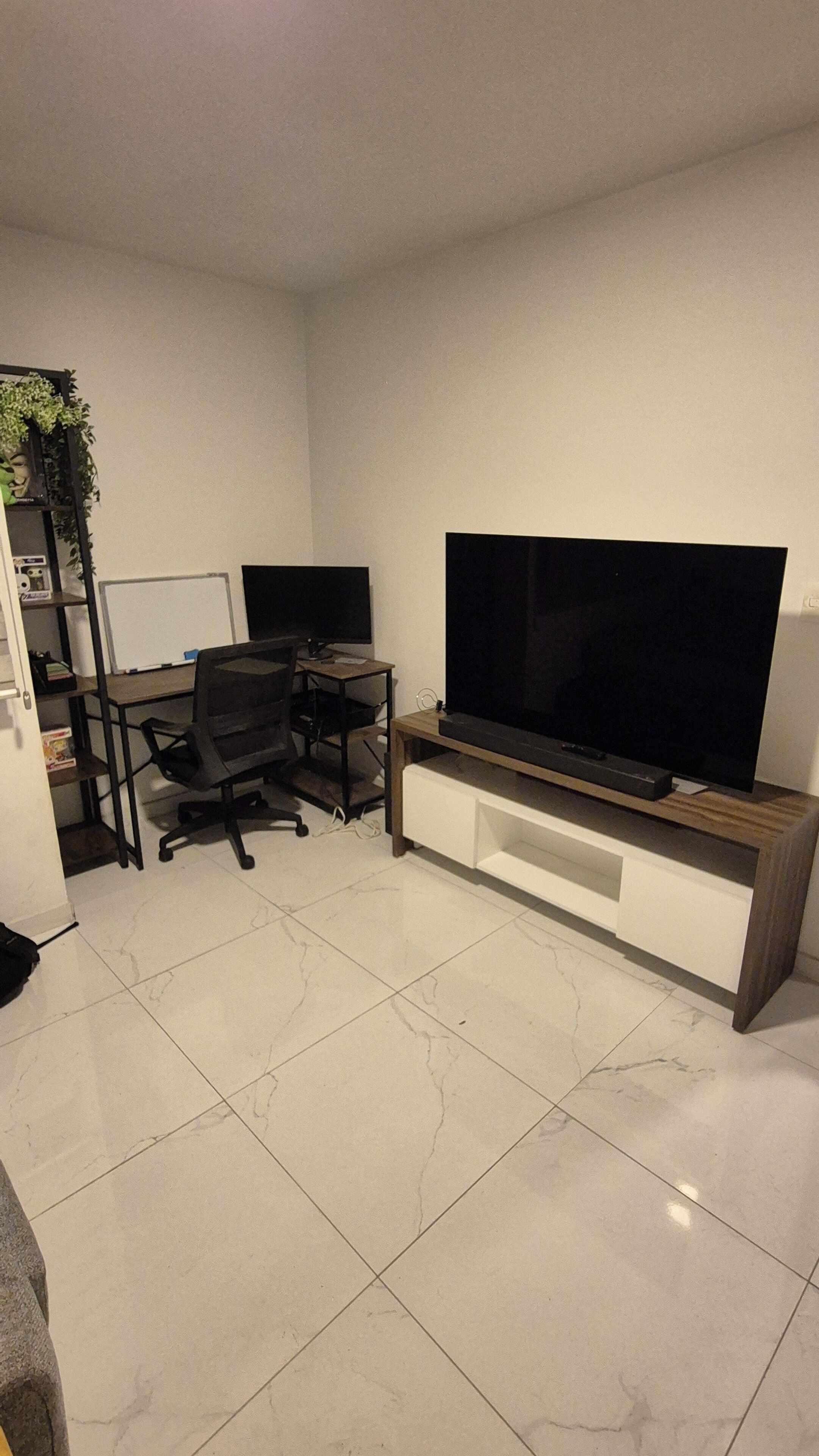 A modern living room with a flat-screen TV, a desk with a chair, and a bookshelf.