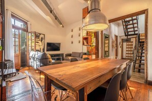 Un vaste espace de vie comprenant une table à manger en bois, un canapé et un téléviseur. Un escalier mène à un autre niveau.
