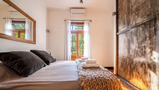 A bedroom with a large bed, a wooden bench, a mirror, and a view of greenery outside. - MOT Rentals + Dorrego House - Pool in Palermo Hollywood (Buenos Aires)