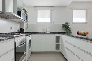 A modern kitchen with white cabinets, a built-in oven, microwave, and a coffee maker. There are two windows providing natural light, and a plant on the countertop. - MOT Rentals + SohoGIam 'B' - 2 Bd 2 Ba (Buenos Aires)