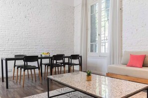 Un coin repas avec un mur en briques blanches, une table avec des chaises, un canapé avec un coussin et une fenêtre avec des stores.