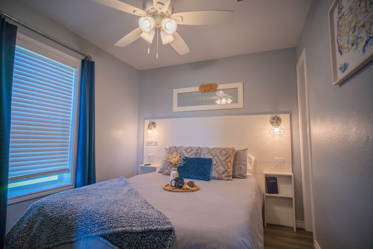 Ein Schlafzimmer mit einem Bett, einem Deckenventilator, einem Nachttisch, einem an der Wand befestigten Regal und einem Fenster mit Jalousien.