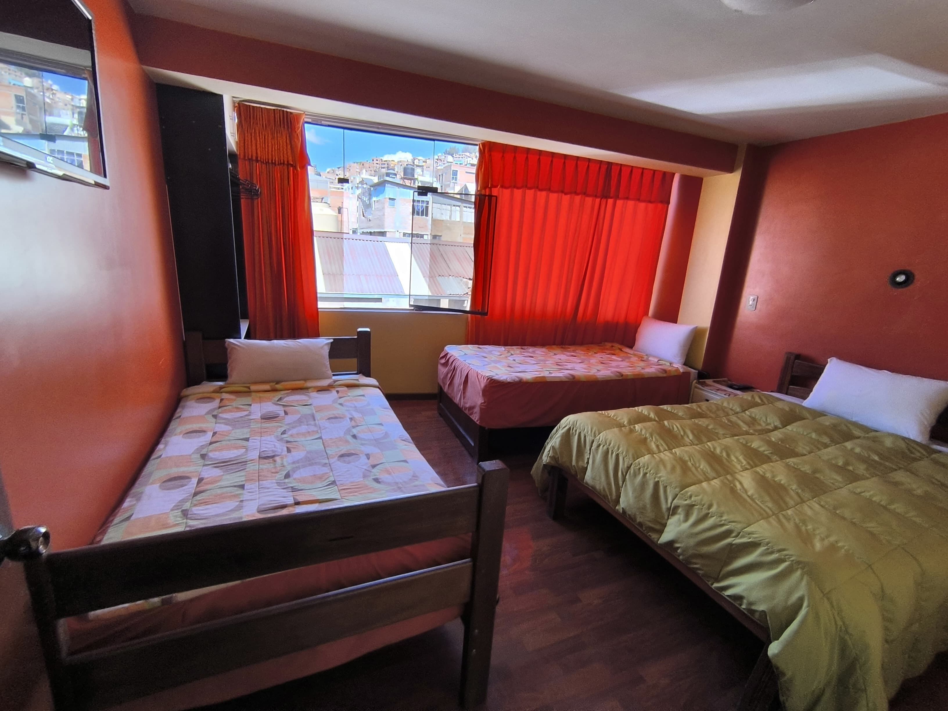 Une chambre avec deux lits, une fenêtre avec des rideaux rouges et une vue sur les bâtiments environnants.