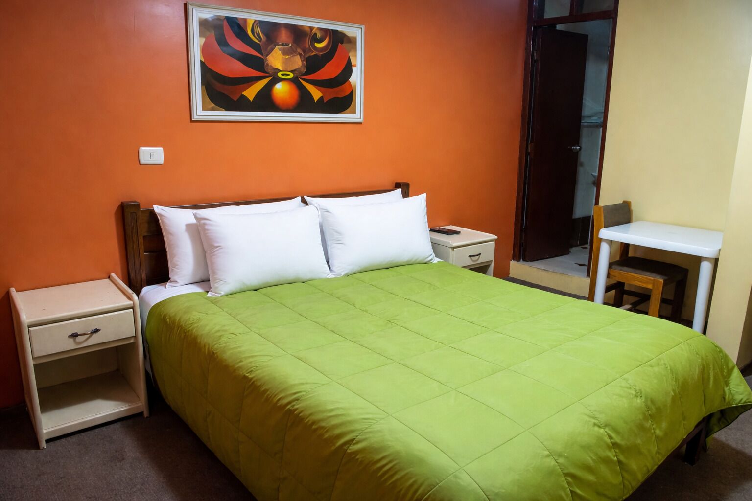 A single-bed room with a green bedspread, white pillows, a nightstand with a drawer, a small table with a chair, and a painting on the wall.