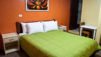 A single-bed room with a green bedspread, white pillows, a nightstand with a drawer, a small table with a chair, and a painting on the wall.
