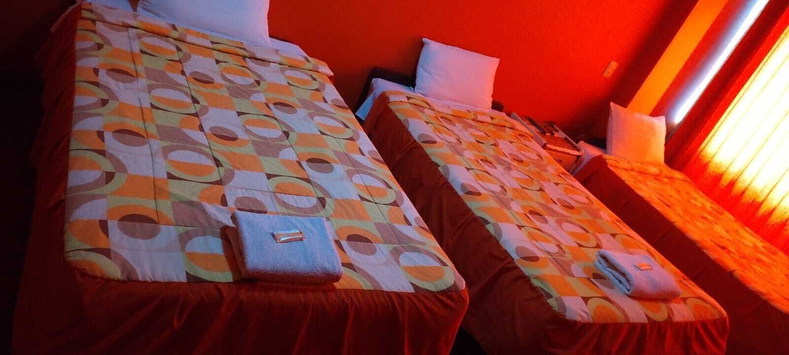 Two beds with patterned bedding, a red wall, and a window with curtains.
