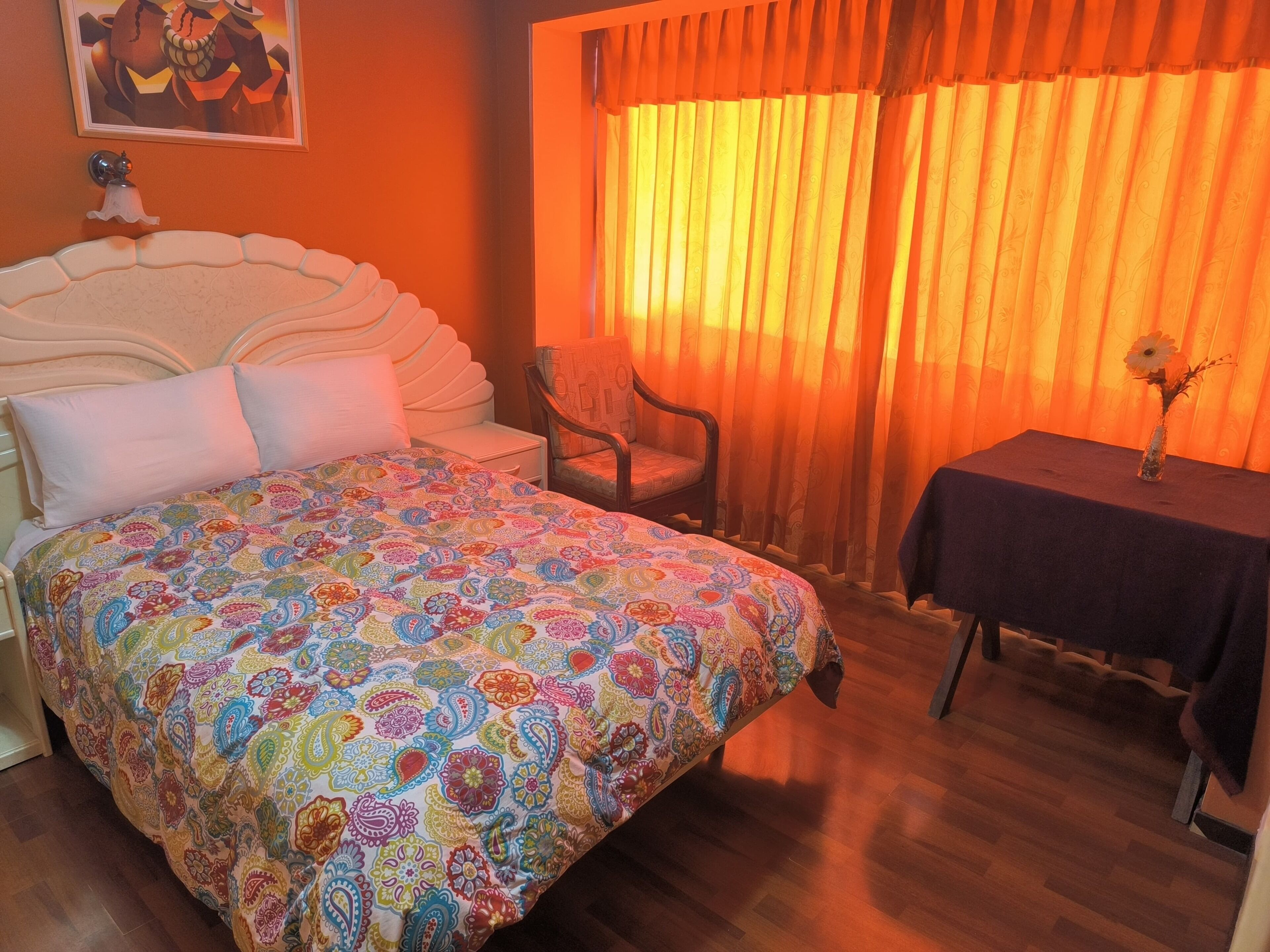 A bed with a colorful patterned bedspread, a chair, a small table with a vase, and a window with orange curtains.