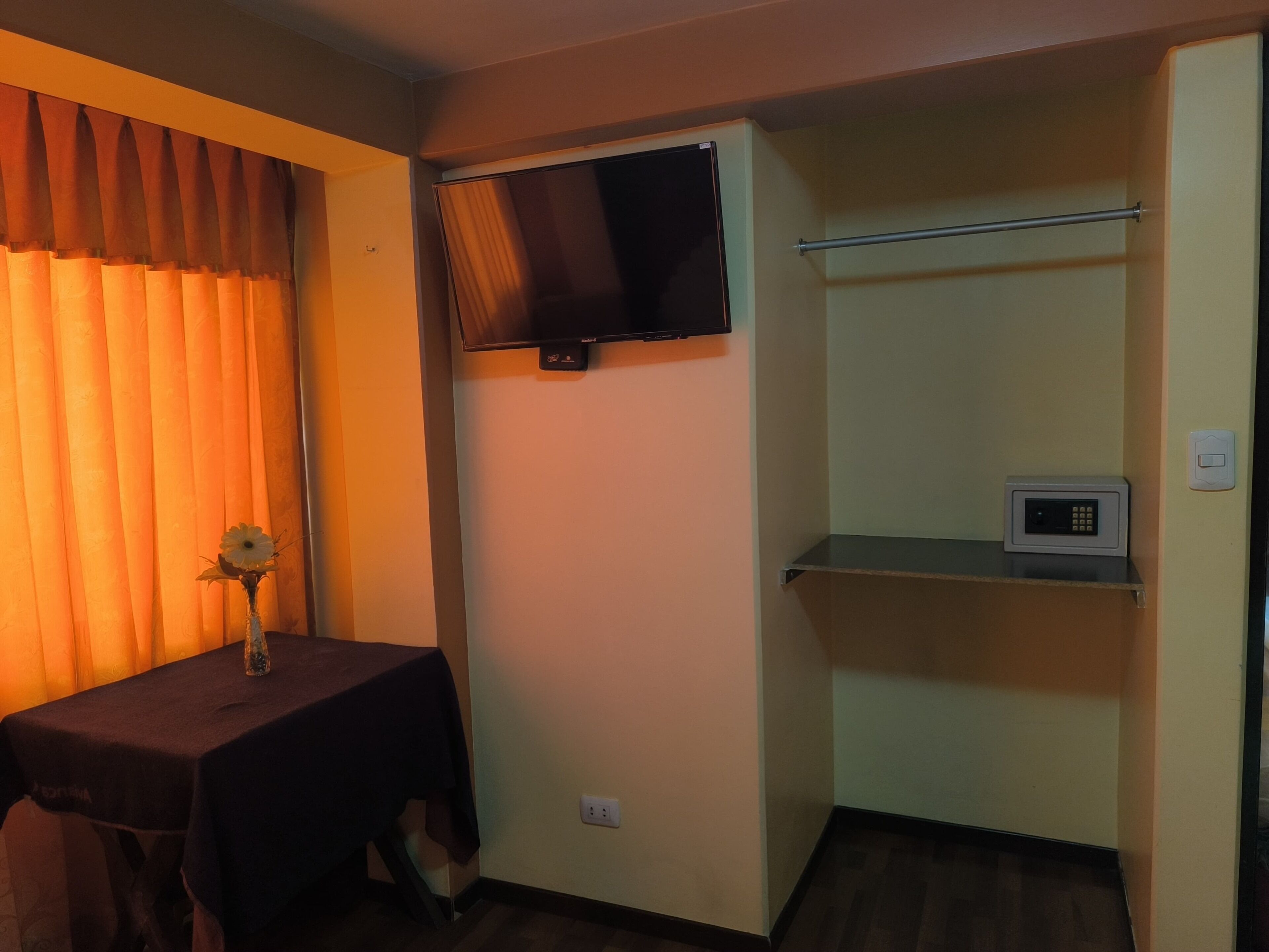 A room with a table, a TV mounted on the wall, and a microwave on a shelf.