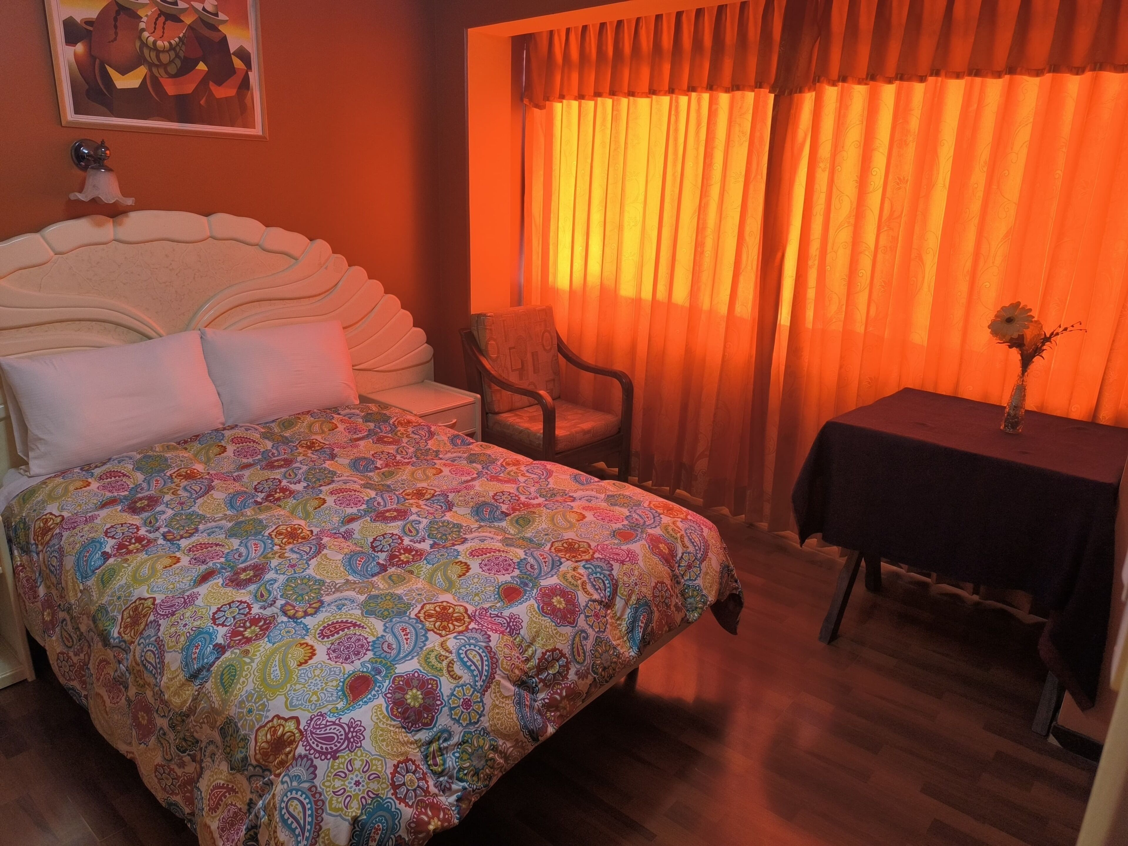 A bedroom with a bed, a chair, a small table with a vase, and a window with curtains.