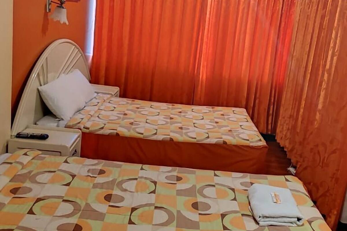 Un letto matrimoniale con copriletto decorato, un comodino con telecomando e tende arancioni.