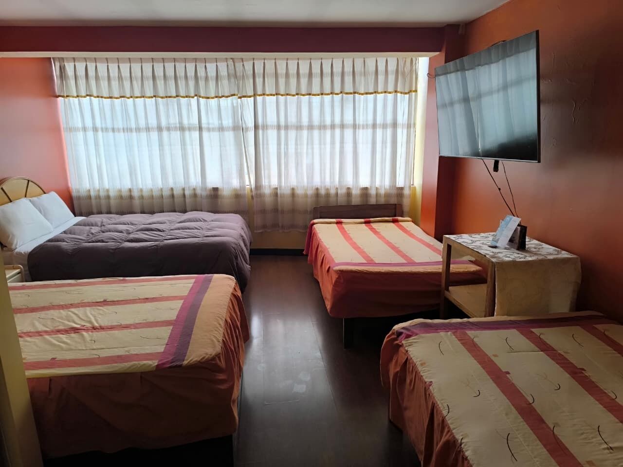 A hotel room with two beds, a TV, a small table with a lamp, and a window with curtains.
