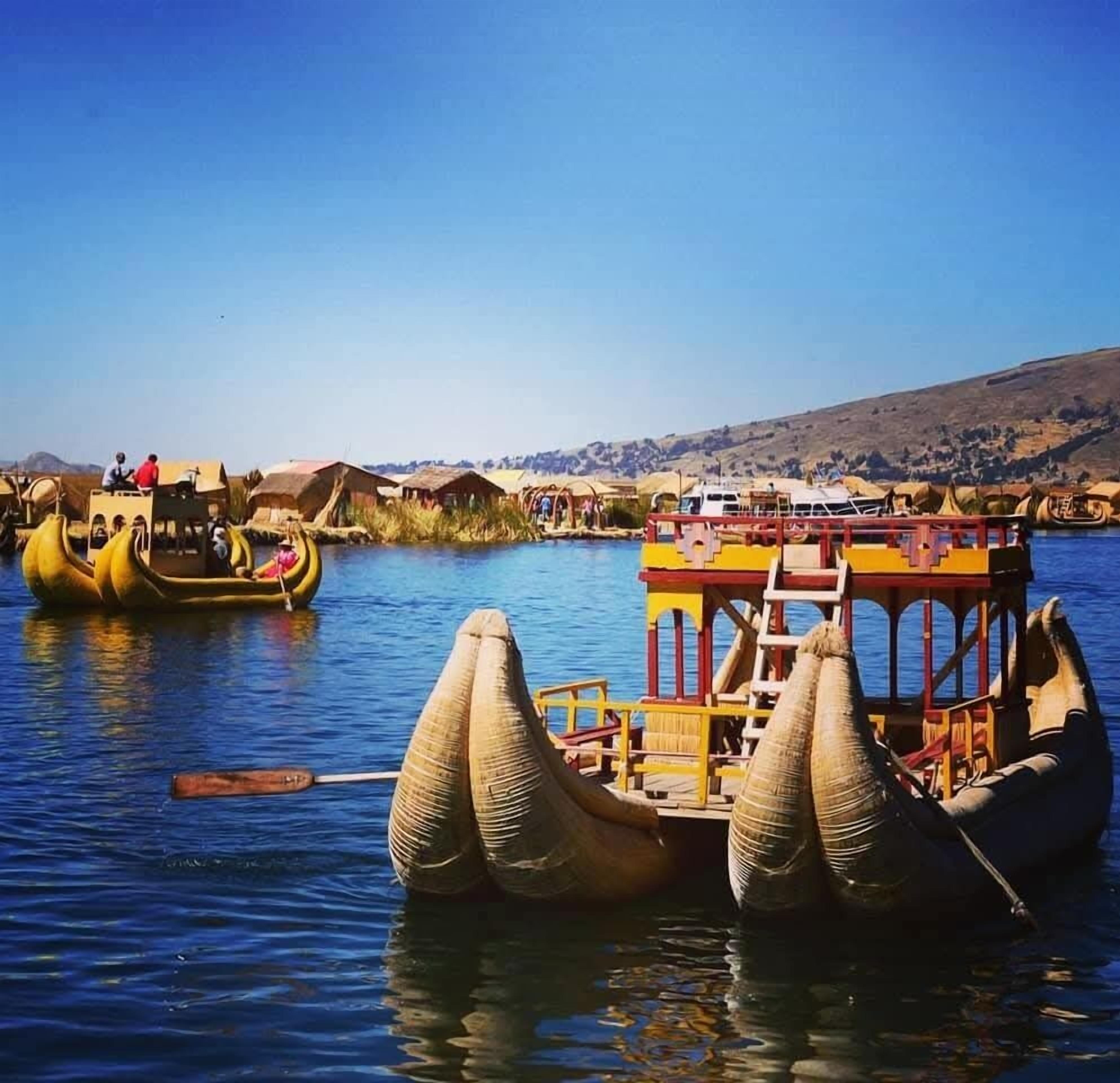 Um barco tradicional com passageiros, um barco atracado e colinas com casas ao fundo.