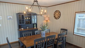 A dining area with a wooden table and chairs, a chandelier, a clock, and a window with blinds. - Lovely 4-bedroom cabin in the heart of Sylvan Lake - 4 min walk to the beach (Sylvan Lake)